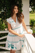 A woman with long brown hair, wearing the Roller Rabbit Avallon Embroidery Pippa Dress, stands on a white porch surrounded by greenery and flowers, gazing thoughtfully into the distance.