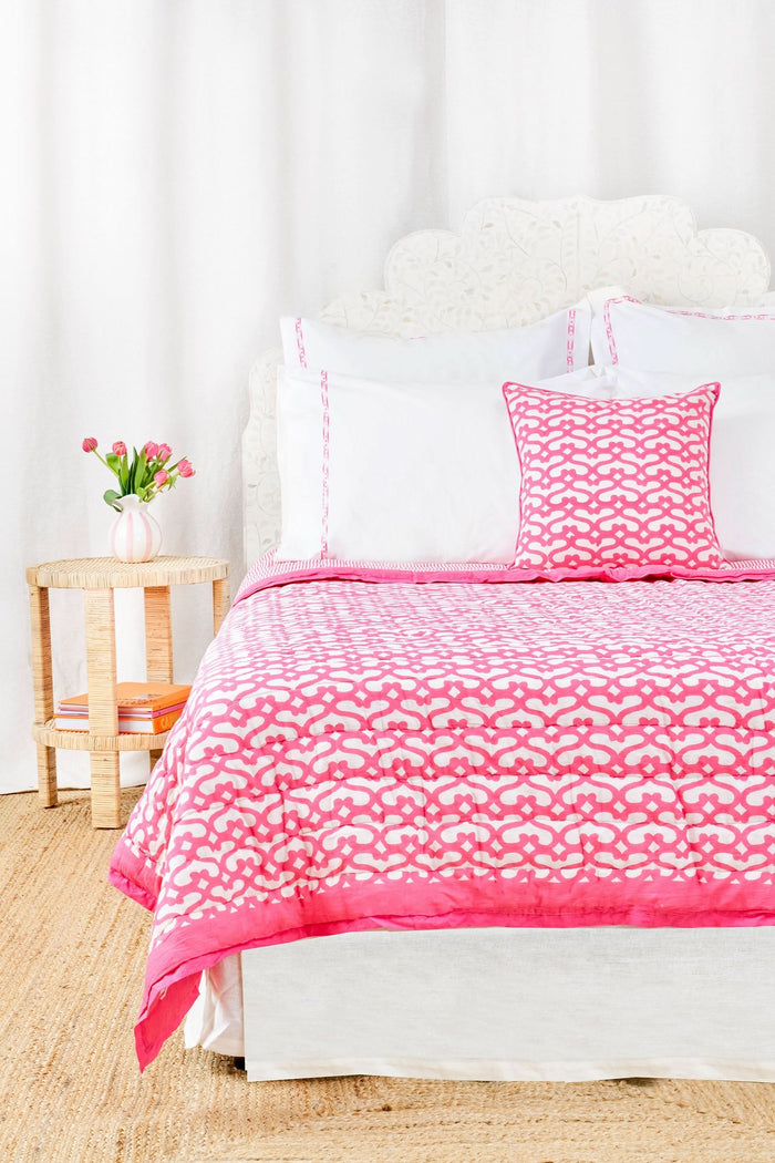 A cozy room features the Roller Rabbit Big Cata Quilt—100 cotton with a white and pink pattern—and a matching pillow on a neatly made bed. A white headboard, side table with pink tulips, books, and white curtains complete the look.