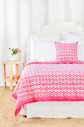 A cozy room features the Roller Rabbit Big Cata Quilt—100 cotton with a white and pink pattern—and a matching pillow on a neatly made bed. A white headboard, side table with pink tulips, books, and white curtains complete the look.