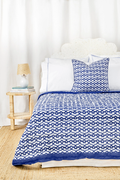 A bed features the Roller Rabbit Big Cata Quilt—white and navy blue with geometric patterns, double-sided—with a matching pillow, white headboard, bedside table with lamp and books, all set before a white curtain backdrop.