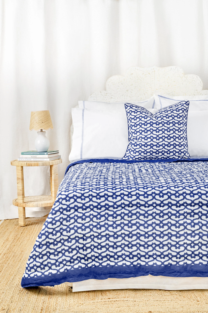 A bed features the Roller Rabbit Big Cata Quilt—white and navy blue with geometric patterns, double-sided—with a matching pillow, white headboard, bedside table with lamp and books, all set before a white curtain backdrop.