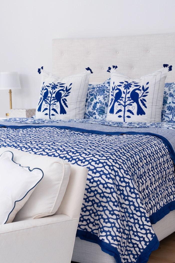 A bed features the Roller Rabbit Big Cata Quilt—white and navy blue with geometric patterns, double-sided—with a matching pillow, white headboard, bedside table with lamp and books, all set before a white curtain backdrop.