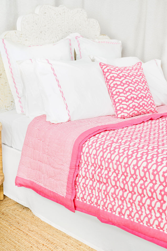 A cozy room features the Roller Rabbit Big Cata Quilt—100 cotton with a white and pink pattern—and a matching pillow on a neatly made bed. A white headboard, side table with pink tulips, books, and white curtains complete the look.