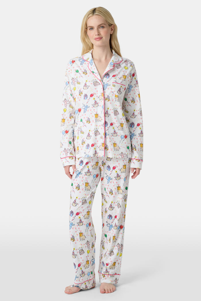 A woman with light skin and long blonde hair wears Roller Rabbit's Birthday Bonanza Long Sleeve Polo Pajamas, a cotton set with colorful cartoon prints and matching button-up top and pants. She stands barefoot with a neutral expression.