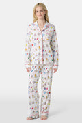 A woman with light skin and long blonde hair wears Roller Rabbit's Birthday Bonanza Long Sleeve Polo Pajamas, a cotton set with colorful cartoon prints and matching button-up top and pants. She stands barefoot with a neutral expression.