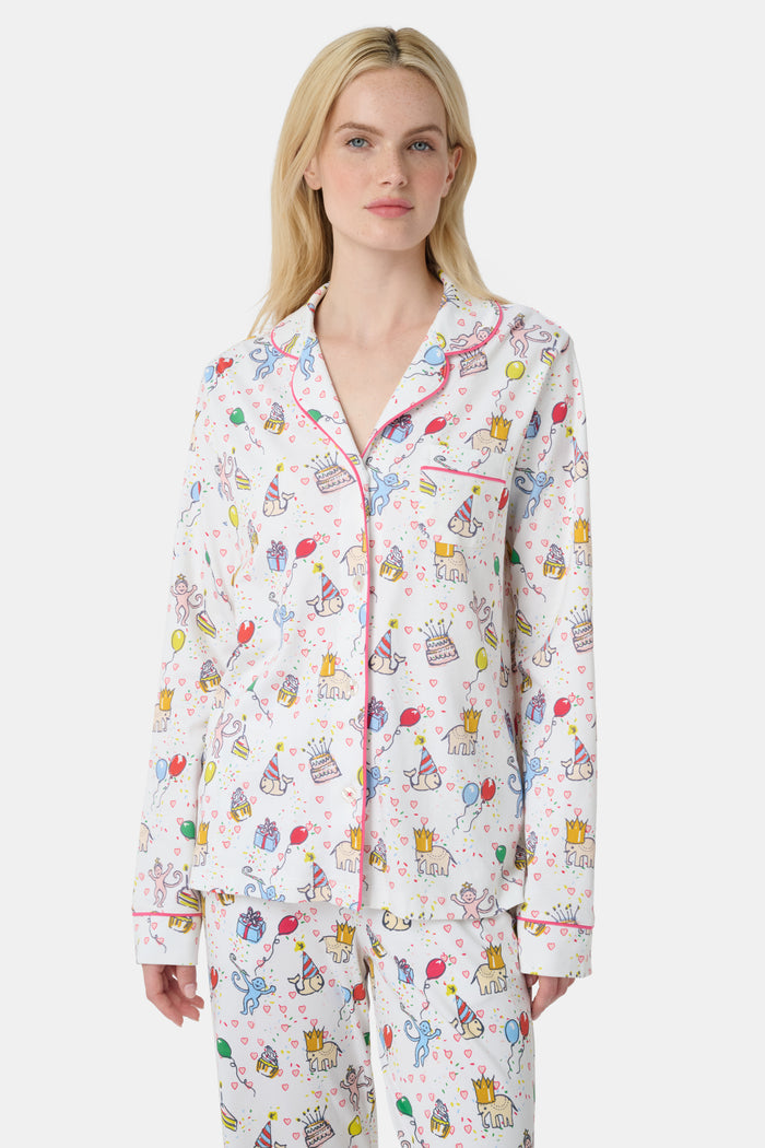 A woman with light skin and long blonde hair wears Roller Rabbit's Birthday Bonanza Long Sleeve Polo Pajamas, a cotton set with colorful cartoon prints and matching button-up top and pants. She stands barefoot with a neutral expression.