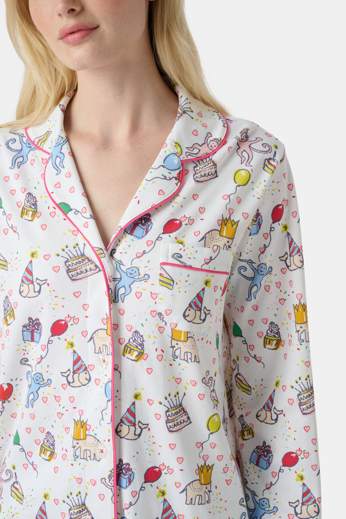 A woman with light skin and long blonde hair wears Roller Rabbit's Birthday Bonanza Long Sleeve Polo Pajamas, a cotton set with colorful cartoon prints and matching button-up top and pants. She stands barefoot with a neutral expression.
