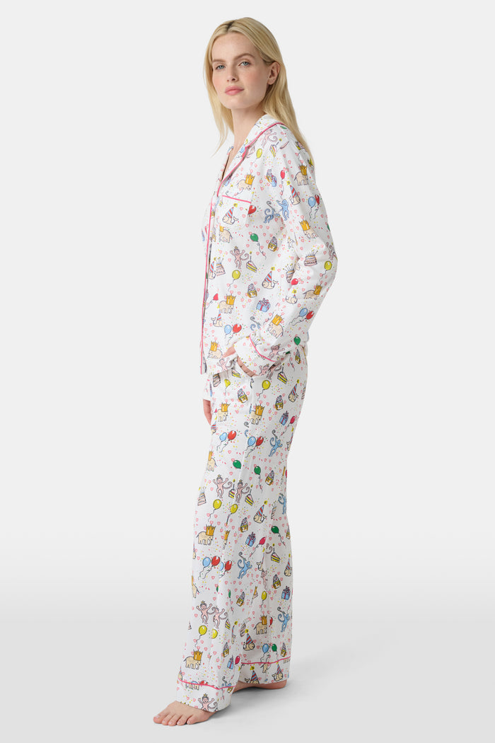 A woman with light skin and long blonde hair wears Roller Rabbit's Birthday Bonanza Long Sleeve Polo Pajamas, a cotton set with colorful cartoon prints and matching button-up top and pants. She stands barefoot with a neutral expression.