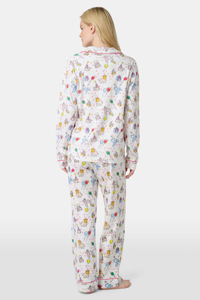 A woman with light skin and long blonde hair wears Roller Rabbit's Birthday Bonanza Long Sleeve Polo Pajamas, a cotton set with colorful cartoon prints and matching button-up top and pants. She stands barefoot with a neutral expression.