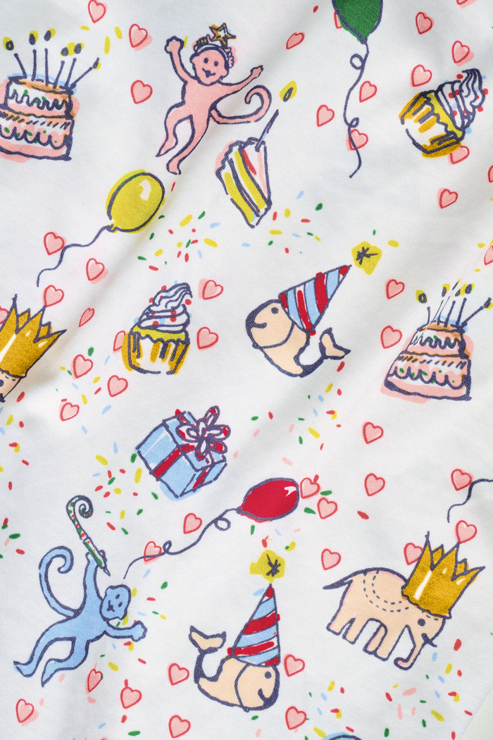 The Roller Rabbit Infant Birthday Bonanza Zipper Footie is a white Pima cotton sleeper with colorful party prints—balloons, cakes, presents, and animals in hats. It has long sleeves and a convenient 2-way zipper for easy changes.
