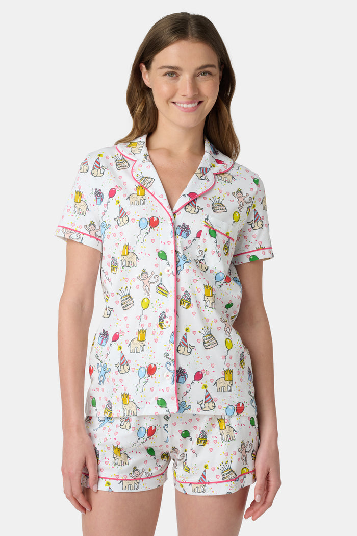 A woman smiles in the Roller Rabbit Birthday Bonanza Polo Pajamas—a women’s short set made from soft Pima cotton with colorful balloon, cake, and animal prints on white.