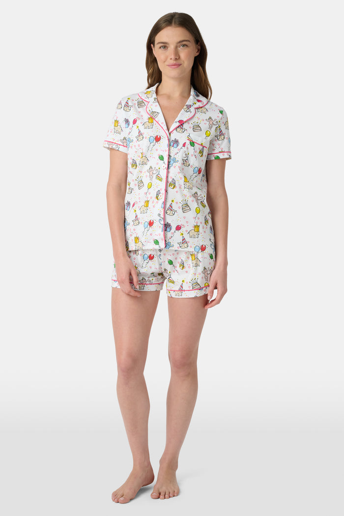 A woman smiles in the Roller Rabbit Birthday Bonanza Polo Pajamas—a women’s short set made from soft Pima cotton with colorful balloon, cake, and animal prints on white.