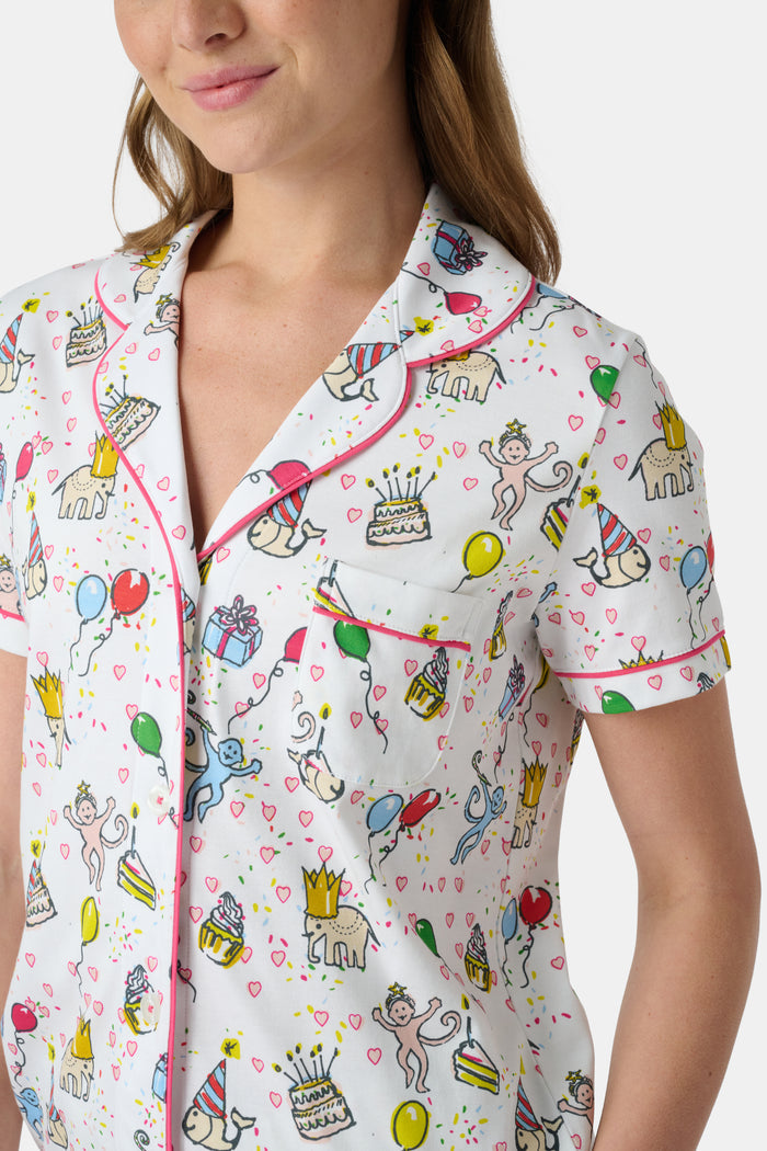 A woman smiles in the Roller Rabbit Birthday Bonanza Polo Pajamas—a women’s short set made from soft Pima cotton with colorful balloon, cake, and animal prints on white.