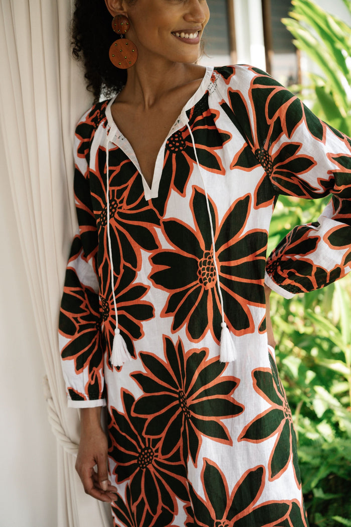 A woman smiles in the Roller Rabbit Blooming Daisies Shiv Caftan, a white maxi dress with bold black and orange daisy prints. She pairs it with orange circular earrings, surrounded by lush green plants in the background.