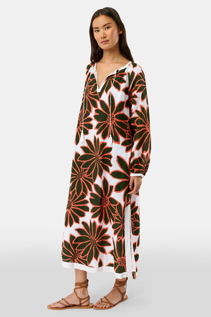 A woman smiles in the Roller Rabbit Blooming Daisies Shiv Caftan, a white maxi dress with bold black and orange daisy prints. She pairs it with orange circular earrings, surrounded by lush green plants in the background.