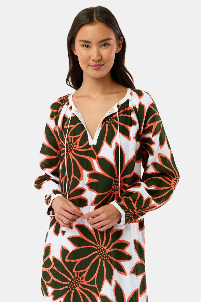 A woman smiles in the Roller Rabbit Blooming Daisies Shiv Caftan, a white maxi dress with bold black and orange daisy prints. She pairs it with orange circular earrings, surrounded by lush green plants in the background.