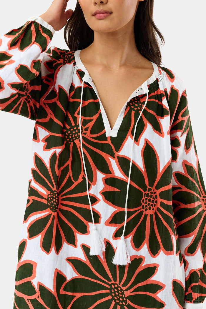 A woman smiles in the Roller Rabbit Blooming Daisies Shiv Caftan, a white maxi dress with bold black and orange daisy prints. She pairs it with orange circular earrings, surrounded by lush green plants in the background.
