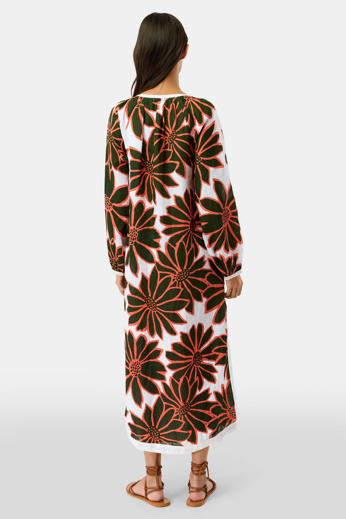 A woman smiles in the Roller Rabbit Blooming Daisies Shiv Caftan, a white maxi dress with bold black and orange daisy prints. She pairs it with orange circular earrings, surrounded by lush green plants in the background.
