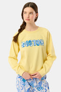 A woman with long brown hair in a loose braid wears the Roller Rabbit Boston Applique Crewneck in light yellow and blue-and-white floral pants, standing against a plain light background.