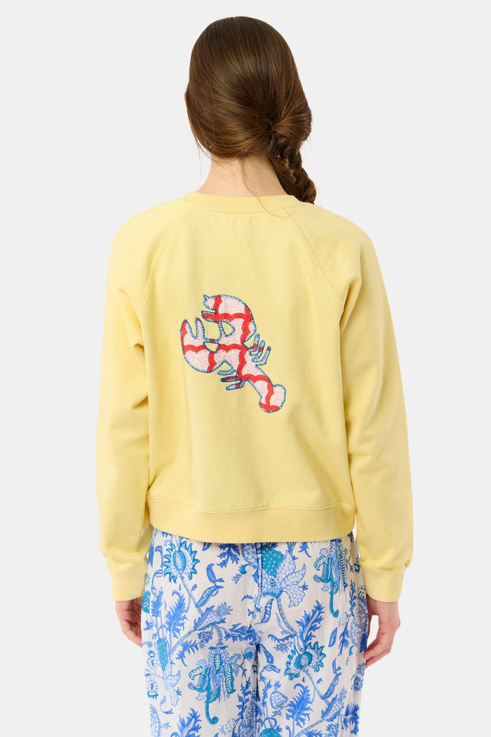 A woman with long brown hair in a loose braid wears the Roller Rabbit Boston Applique Crewneck in light yellow and blue-and-white floral pants, standing against a plain light background.