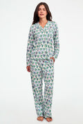 A child stands barefoot against a plain white background, wearing the Roller Rabbit Boston Monkey Long Sleeve Polo Pajamas—matching white cotton sleepwear adorned with a colorful floral and animal print. These soft children’s pajamas feature long sleeves and full-length pants for cozy bedtime comfort and playful style.