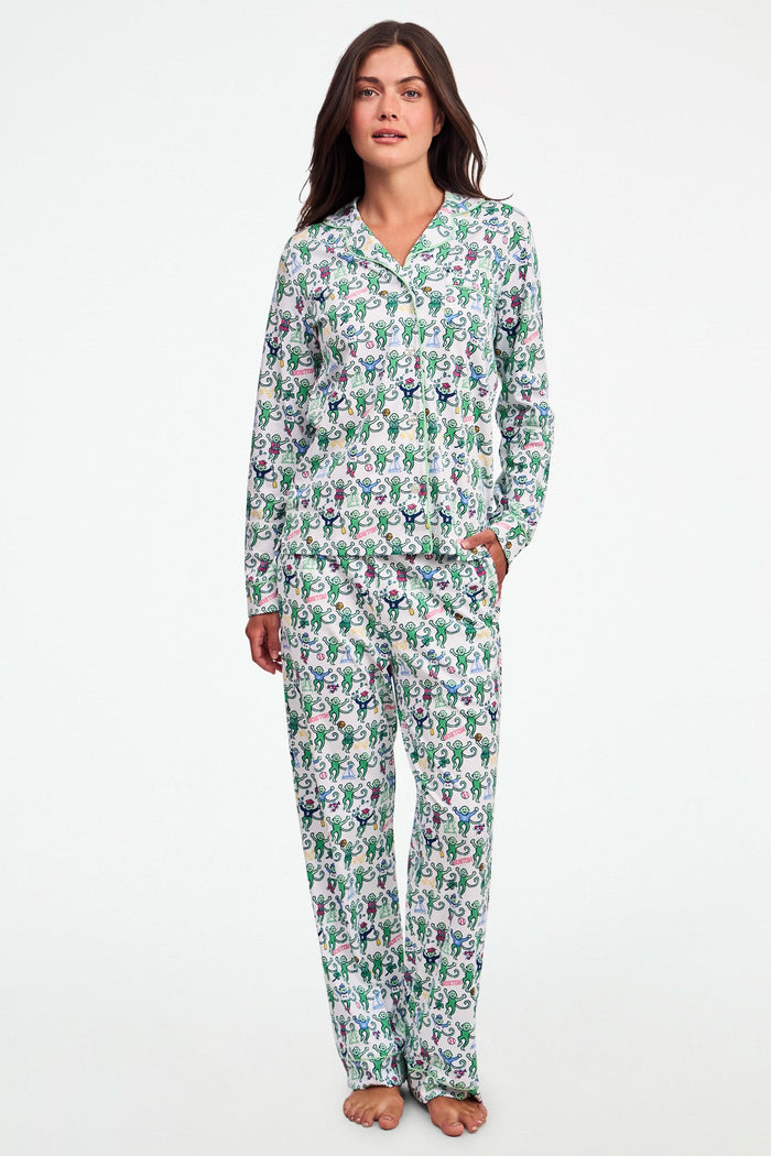 A child stands barefoot against a plain white background, wearing the Roller Rabbit Boston Monkey Long Sleeve Polo Pajamas—matching white cotton sleepwear adorned with a colorful floral and animal print. These soft children’s pajamas feature long sleeves and full-length pants for cozy bedtime comfort and playful style.