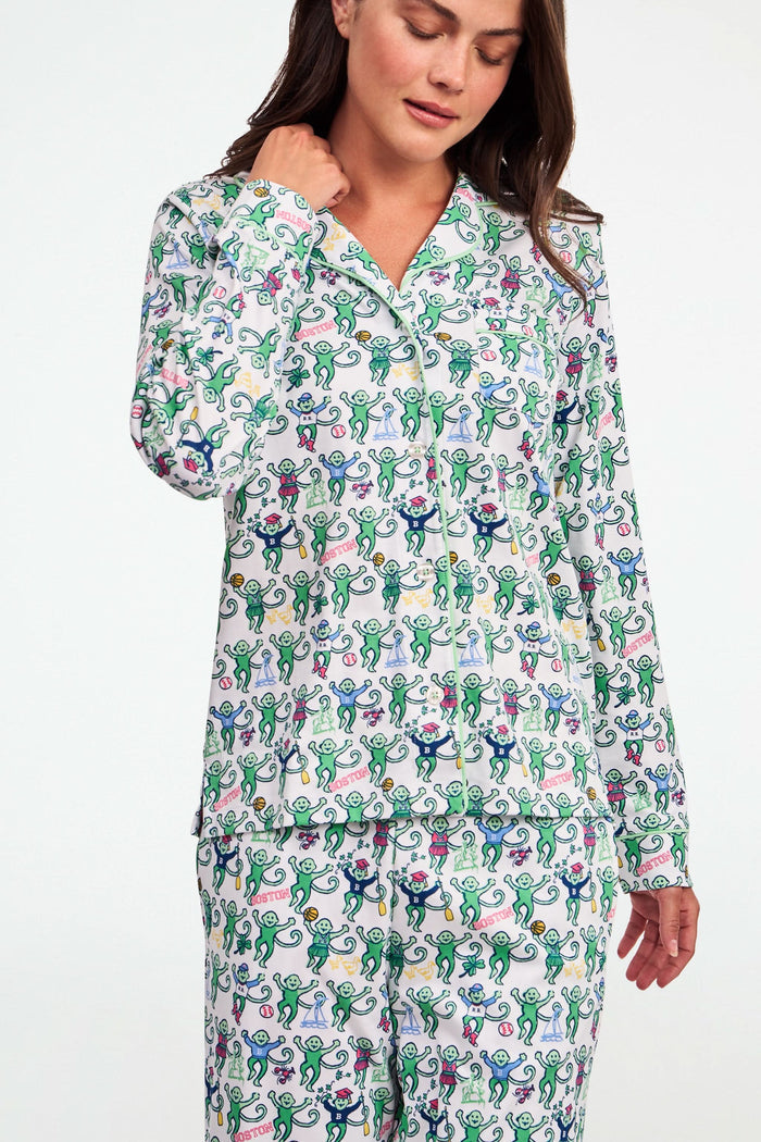 A child stands barefoot against a plain white background, wearing the Roller Rabbit Boston Monkey Long Sleeve Polo Pajamas—matching white cotton sleepwear adorned with a colorful floral and animal print. These soft children’s pajamas feature long sleeves and full-length pants for cozy bedtime comfort and playful style.