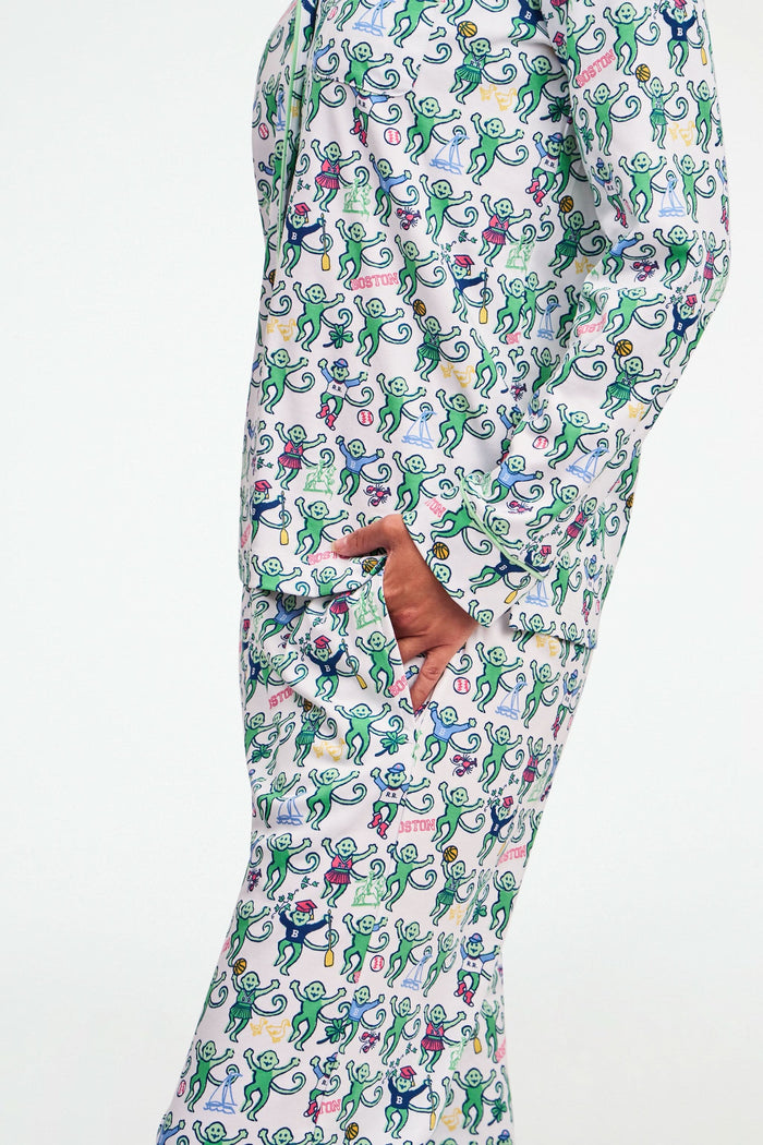 A child stands barefoot against a plain white background, wearing the Roller Rabbit Boston Monkey Long Sleeve Polo Pajamas—matching white cotton sleepwear adorned with a colorful floral and animal print. These soft children’s pajamas feature long sleeves and full-length pants for cozy bedtime comfort and playful style.