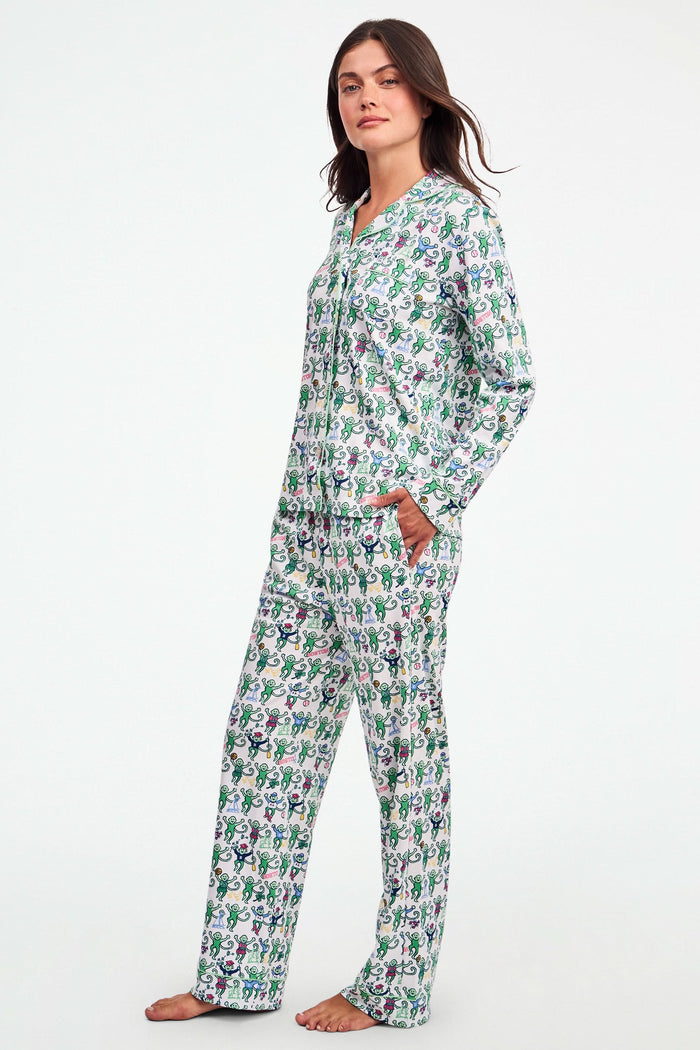 A child stands barefoot against a plain white background, wearing the Roller Rabbit Boston Monkey Long Sleeve Polo Pajamas—matching white cotton sleepwear adorned with a colorful floral and animal print. These soft children’s pajamas feature long sleeves and full-length pants for cozy bedtime comfort and playful style.