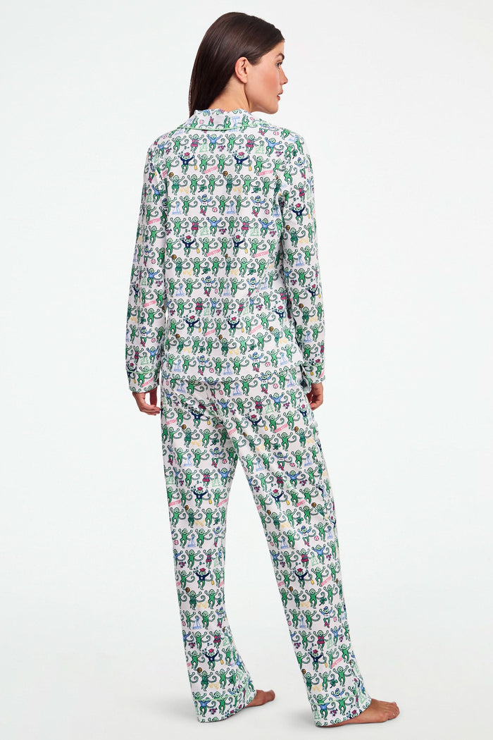 A child stands barefoot against a plain white background, wearing the Roller Rabbit Boston Monkey Long Sleeve Polo Pajamas—matching white cotton sleepwear adorned with a colorful floral and animal print. These soft children’s pajamas feature long sleeves and full-length pants for cozy bedtime comfort and playful style.