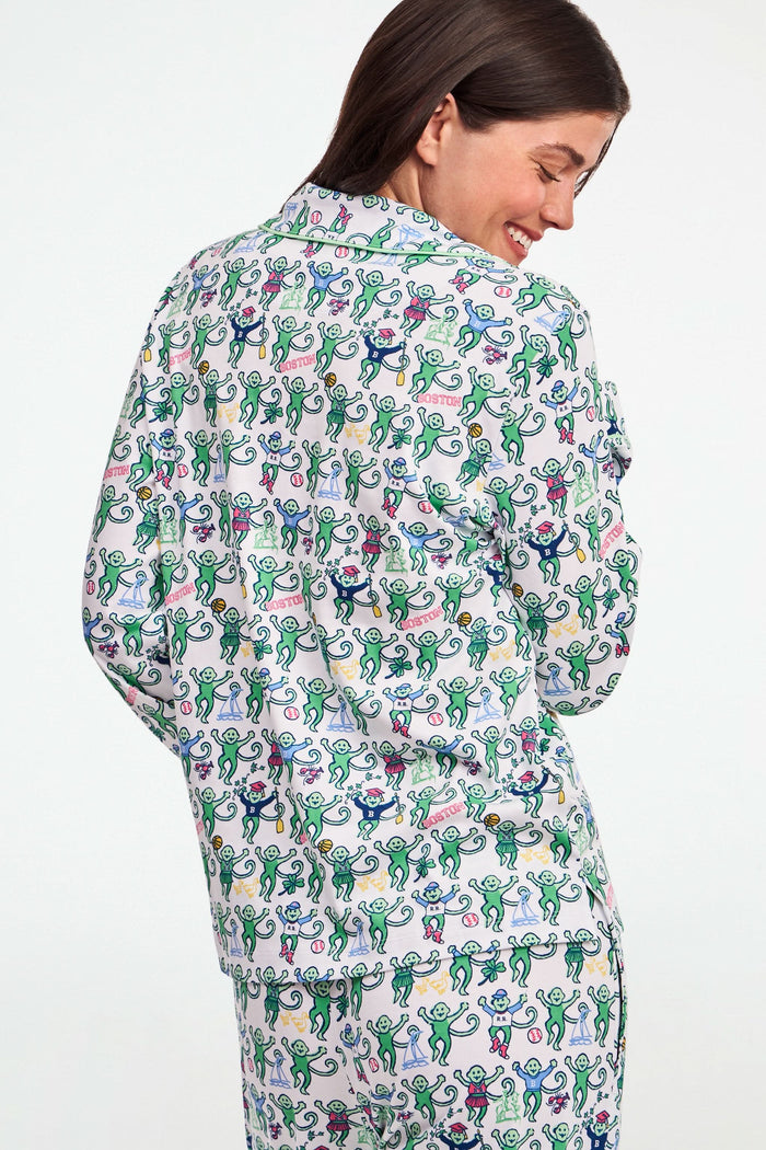 A child stands barefoot against a plain white background, wearing the Roller Rabbit Boston Monkey Long Sleeve Polo Pajamas—matching white cotton sleepwear adorned with a colorful floral and animal print. These soft children’s pajamas feature long sleeves and full-length pants for cozy bedtime comfort and playful style.
