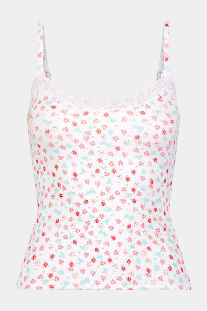 A woman with long dark hair wears the Roller Rabbit Bouquet Pointelle Carmine Cami, featuring pink and blue floral print and lace trim. She smiles softly as she looks down, standing against a plain light background.