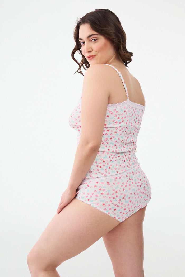A woman with long dark hair wears the Roller Rabbit Bouquet Pointelle Carmine Cami, featuring pink and blue floral print and lace trim. She smiles softly as she looks down, standing against a plain light background.