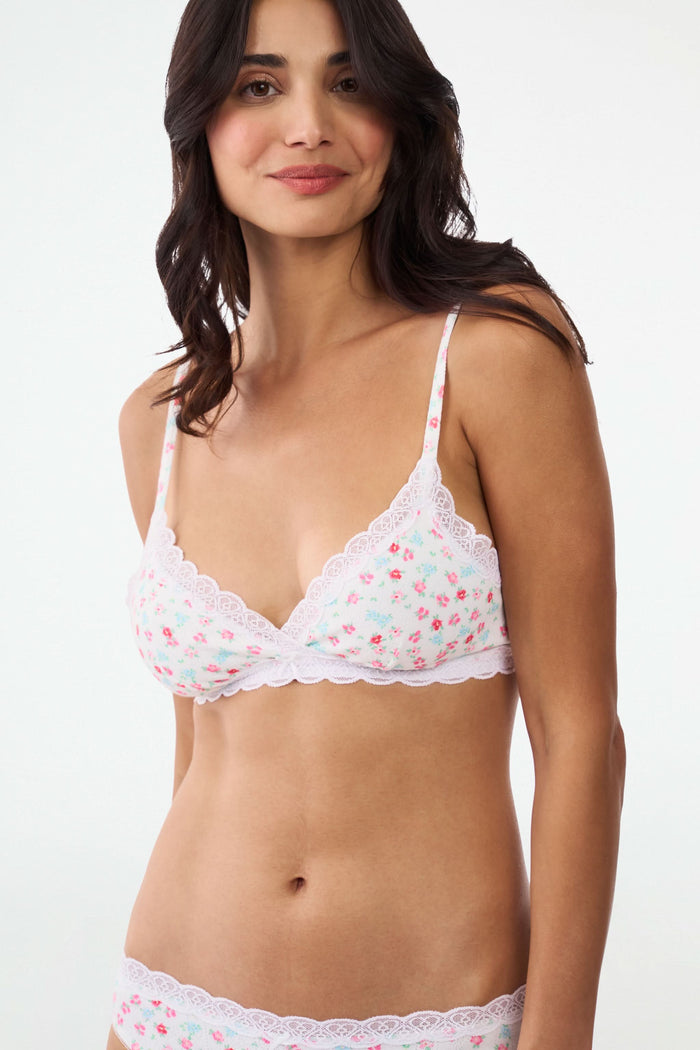 A woman with wavy brown hair smiles with one arm raised, wearing the Roller Rabbit Bouquet Pointelle Jane Bralette and matching underwear against a plain white background.