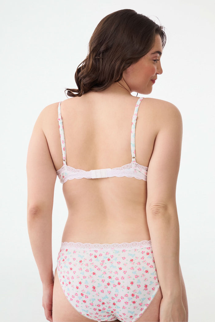 A woman with wavy brown hair smiles with one arm raised, wearing the Roller Rabbit Bouquet Pointelle Jane Bralette and matching underwear against a plain white background.