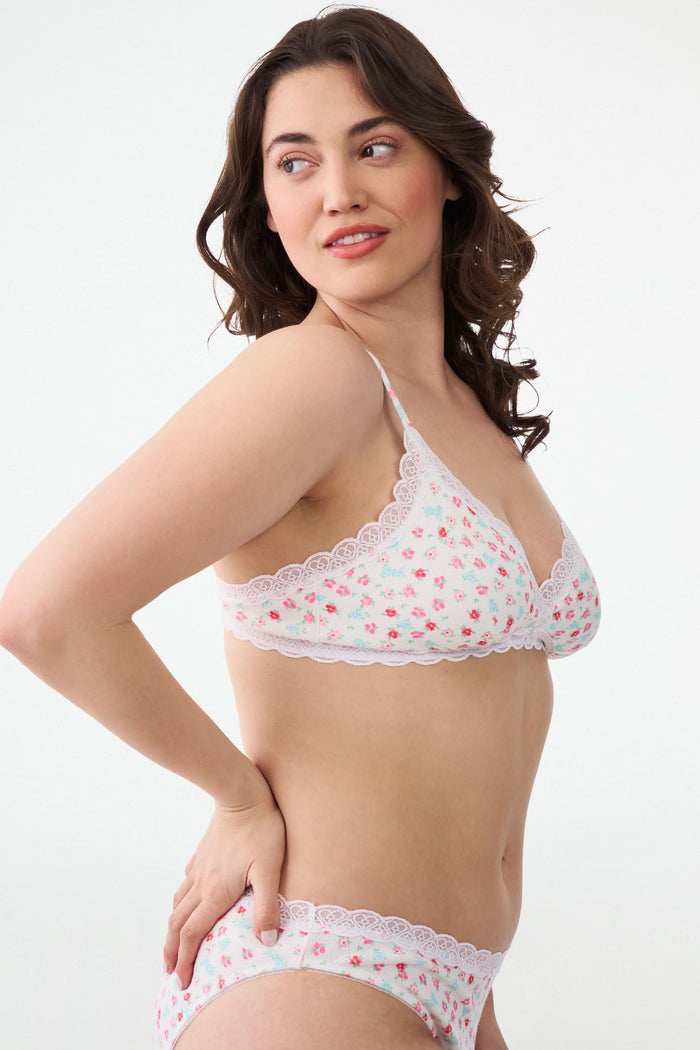 A woman with wavy brown hair smiles with one arm raised, wearing the Roller Rabbit Bouquet Pointelle Jane Bralette and matching underwear against a plain white background.