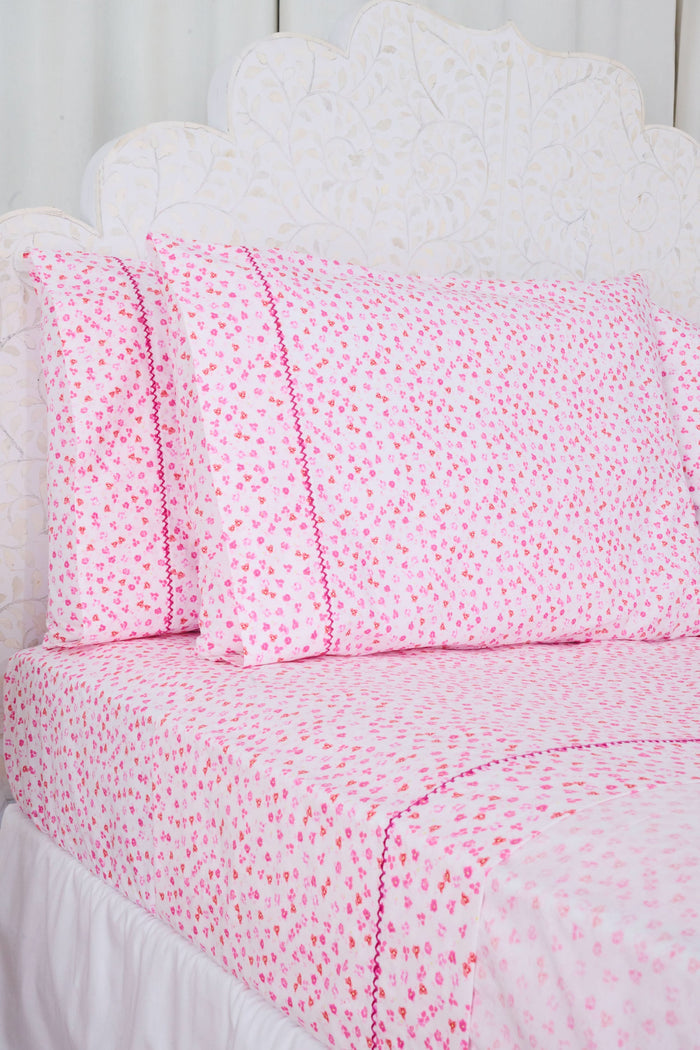 The bed features Roller Rabbit’s Bouquet Sheet Set, with a white ornate headboard and light pink 300 thread count cotton sheets and pillowcases adorned with small hearts and decorative pink stitching for a soft, cozy look.