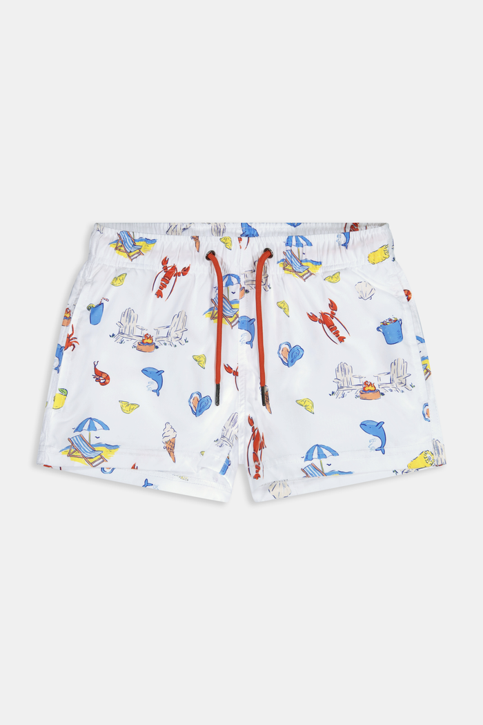 A young boy smiles barefoot in front of a cream-colored vehicle, wearing Roller Rabbit Boys Catch of the Day Shortie Swim Trunk with colorful prints, while another child sits partially visible beside him.