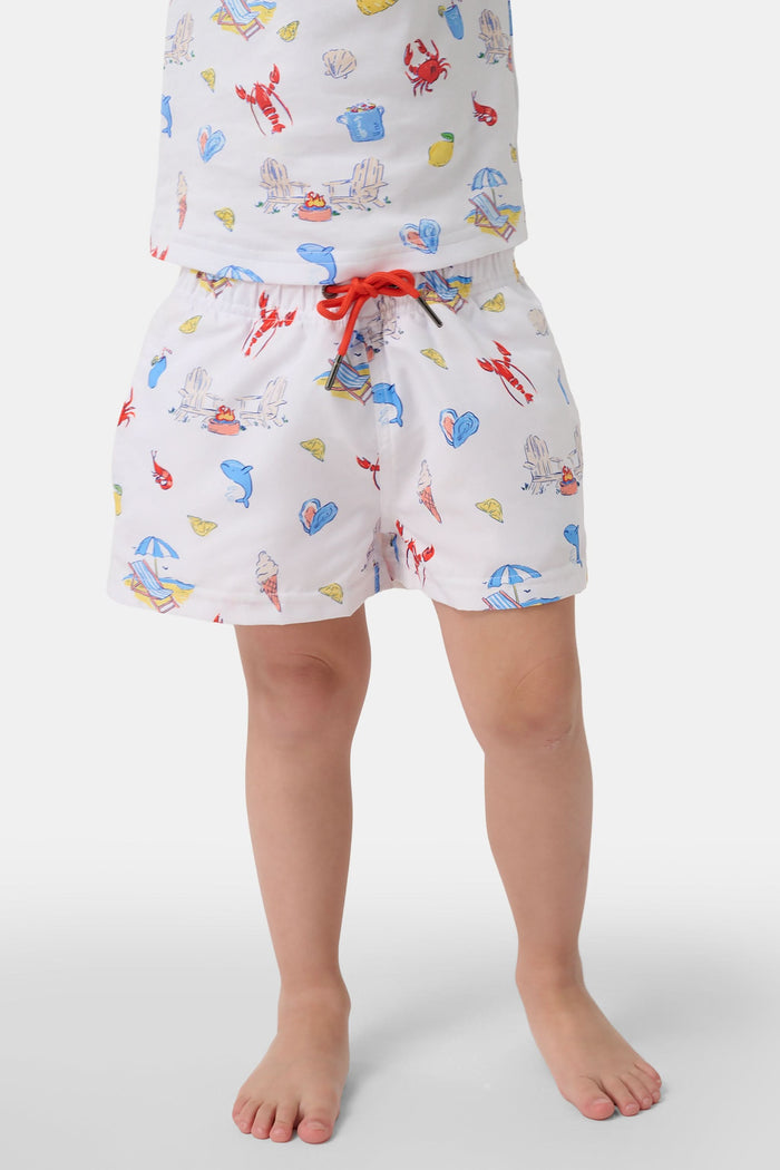 A young boy smiles barefoot in front of a cream-colored vehicle, wearing Roller Rabbit Boys Catch of the Day Shortie Swim Trunk with colorful prints, while another child sits partially visible beside him.
