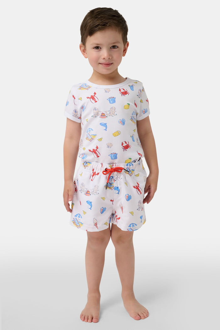 A young boy smiles barefoot in front of a cream-colored vehicle, wearing Roller Rabbit Boys Catch of the Day Shortie Swim Trunk with colorful prints, while another child sits partially visible beside him.