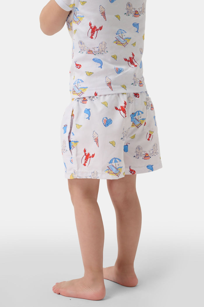 A young boy smiles barefoot in front of a cream-colored vehicle, wearing Roller Rabbit Boys Catch of the Day Shortie Swim Trunk with colorful prints, while another child sits partially visible beside him.