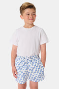 A young boy with light hair wears a plain white T-shirt and Roller Rabbit Boys Moby Swim Trunk. He stands against a white background, one hand in his pocket, smiling slightly and looking to the side.