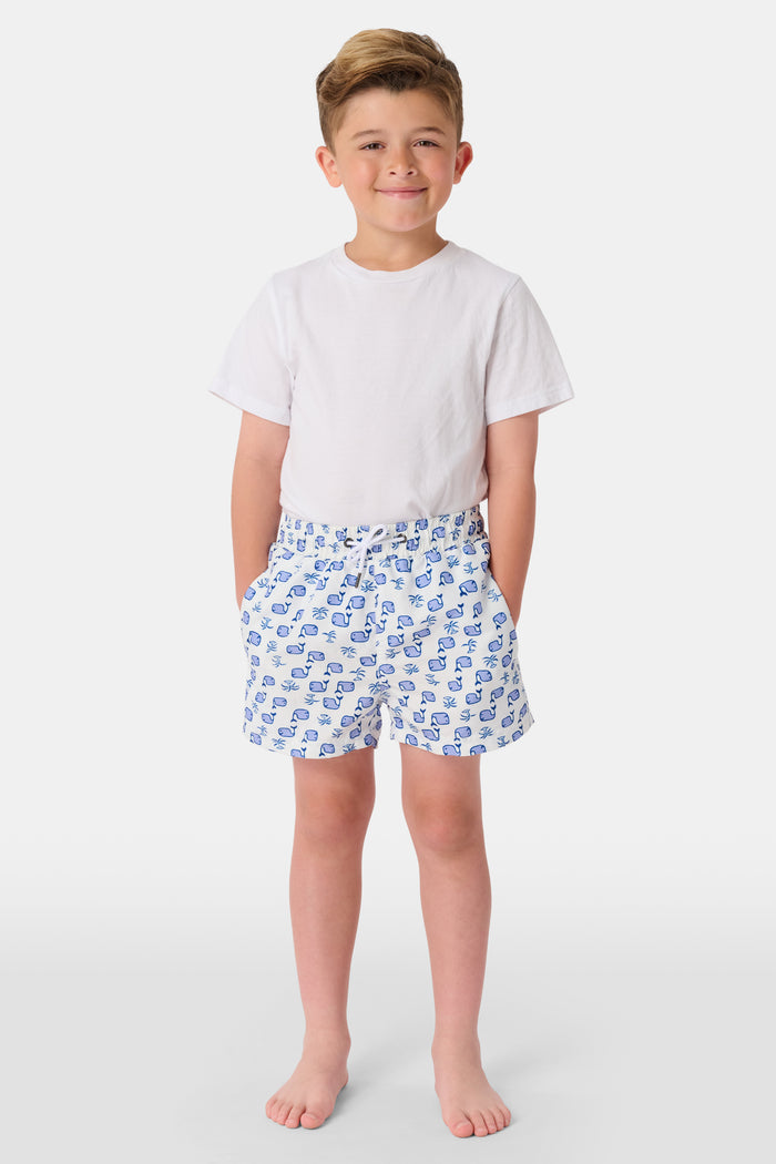 A young boy with light hair wears a plain white T-shirt and Roller Rabbit Boys Moby Swim Trunk. He stands against a white background, one hand in his pocket, smiling slightly and looking to the side.