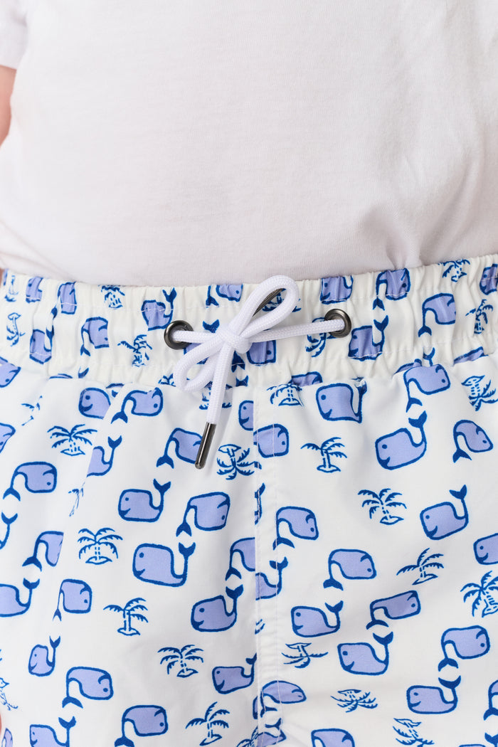 A young boy with light hair wears a plain white T-shirt and Roller Rabbit Boys Moby Swim Trunk. He stands against a white background, one hand in his pocket, smiling slightly and looking to the side.