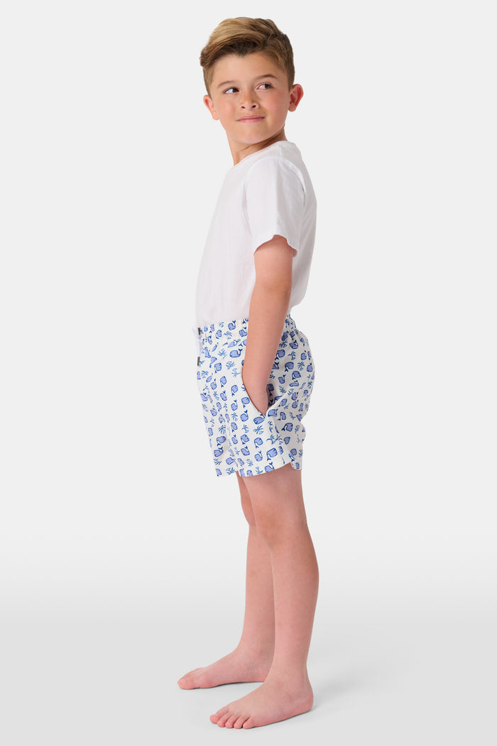 A young boy with light hair wears a plain white T-shirt and Roller Rabbit Boys Moby Swim Trunk. He stands against a white background, one hand in his pocket, smiling slightly and looking to the side.