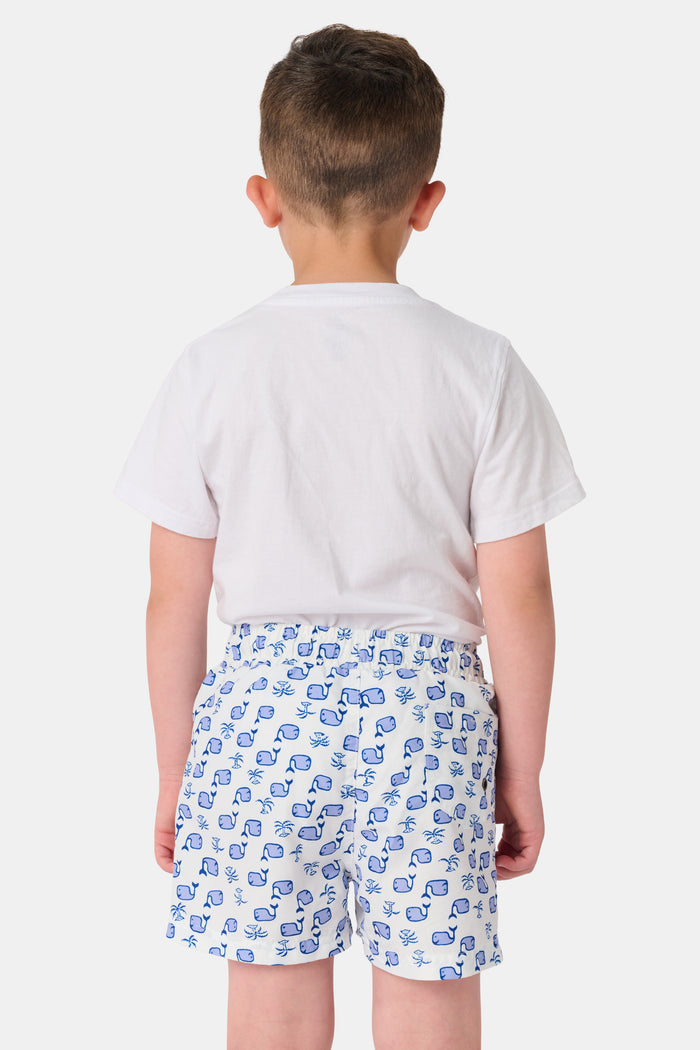 A young boy with light hair wears a plain white T-shirt and Roller Rabbit Boys Moby Swim Trunk. He stands against a white background, one hand in his pocket, smiling slightly and looking to the side.