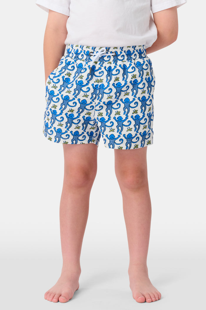 A child stands barefoot in a white t-shirt and Roller Rabbit Boys Monkey Swim Trunks, featuring a playful blue monkey print, with hands in pockets against a plain white background.
