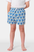 A child stands barefoot in a white t-shirt and Roller Rabbit Boys Monkey Swim Trunks, featuring a playful blue monkey print, with hands in pockets against a plain white background.