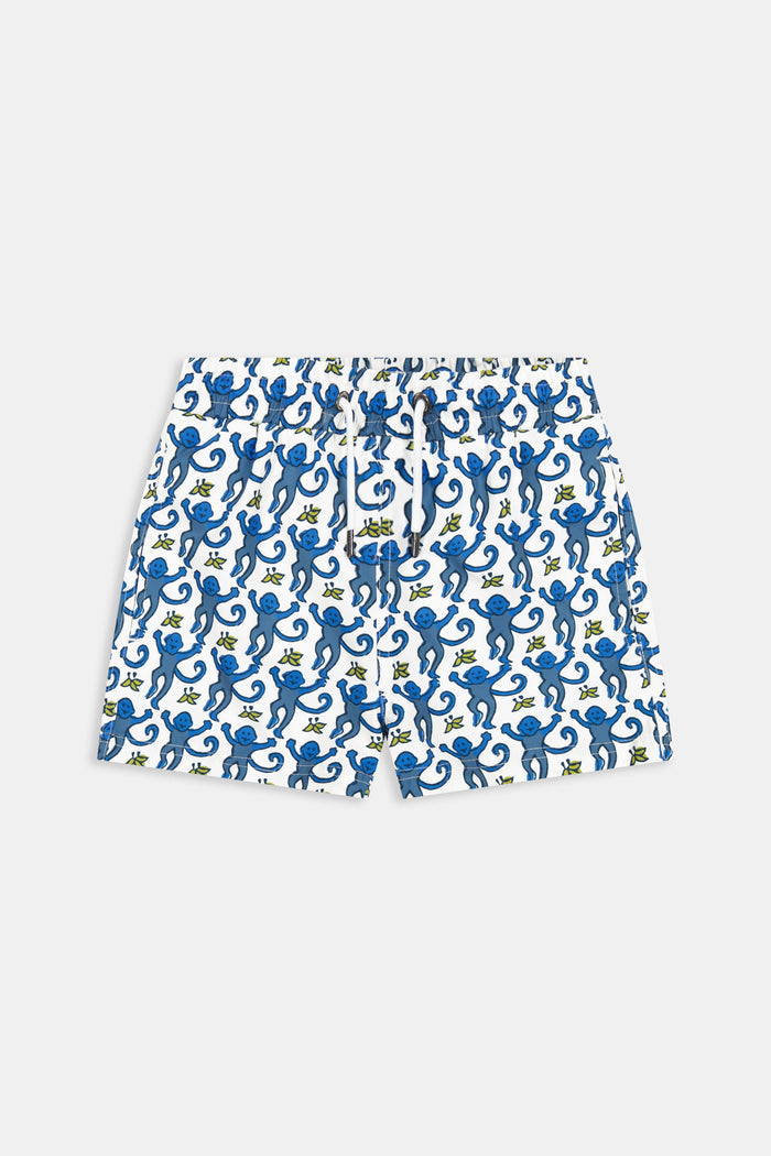 A child stands barefoot in a white t-shirt and Roller Rabbit Boys Monkey Swim Trunks, featuring a playful blue monkey print, with hands in pockets against a plain white background.
