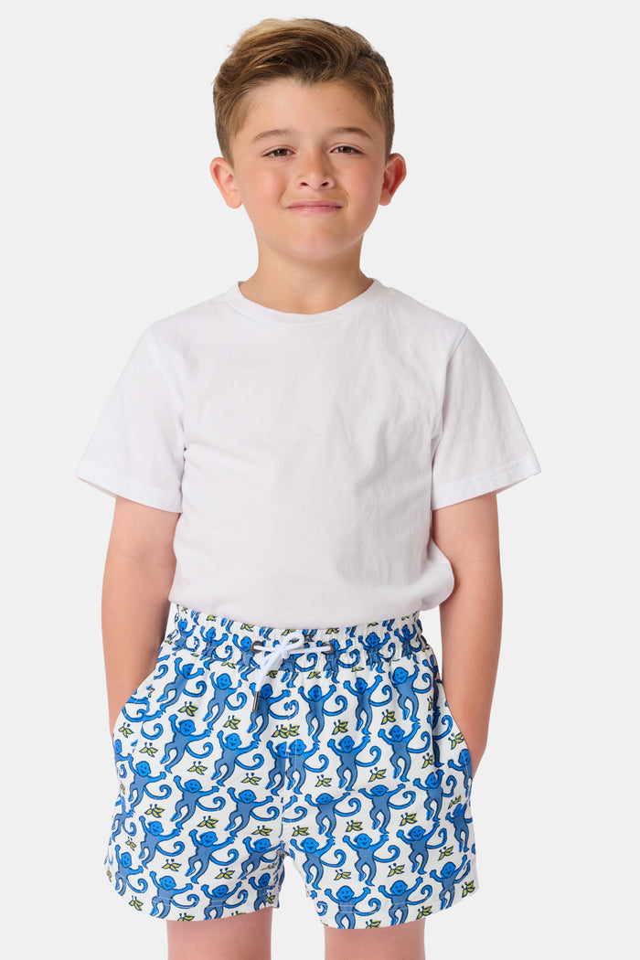 A child stands barefoot in a white t-shirt and Roller Rabbit Boys Monkey Swim Trunks, featuring a playful blue monkey print, with hands in pockets against a plain white background.